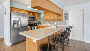 A modern kitchen with a stainless steel refrigerator, wooden cabinets, a large island with bar stools, and a white door. - Gulf Coast Get-Away! Heated pool, sleeps 10! 69th (Largo)