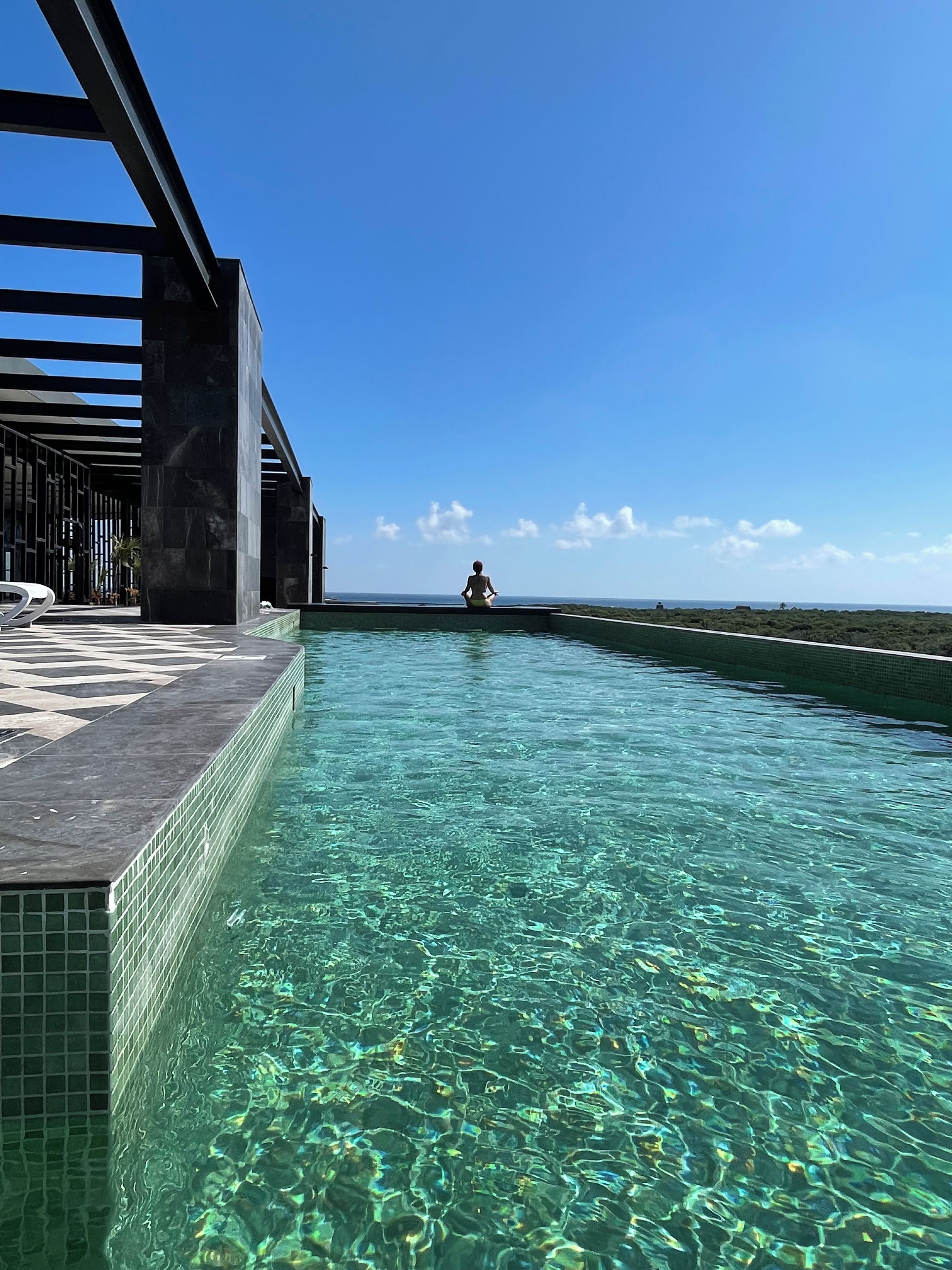 A clear pool with a person in the distance, surrounded by a modern structure with a tiled floor.