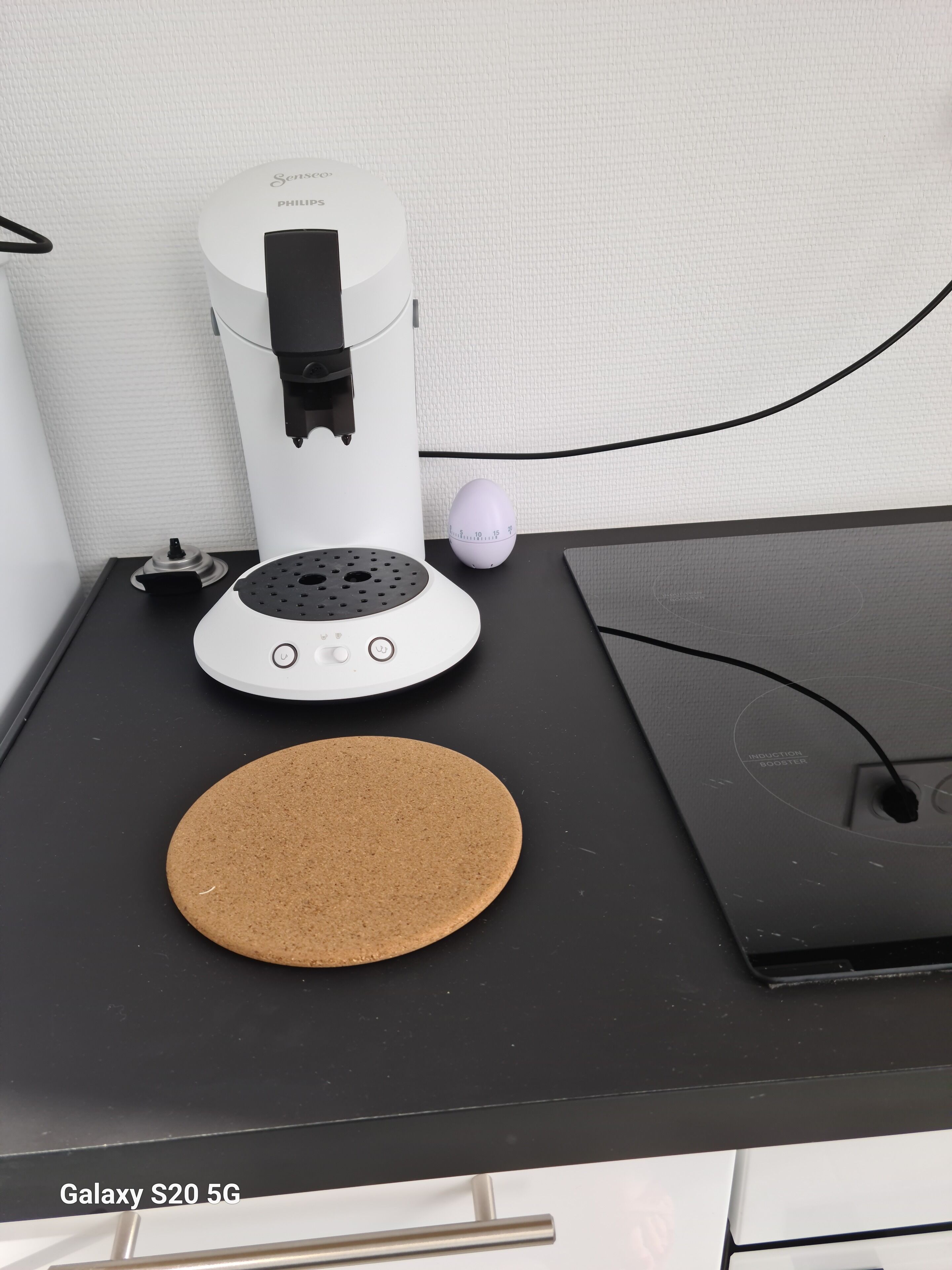 Un smartphone Galaxy S20 5G posé sur une surface noire, avec une machine à café Philips blanche, un minuteur à œufs violet et un sous-verre en liège.