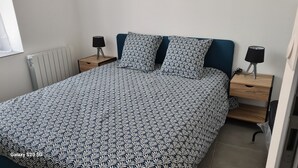 Ein ordentlich bezogenes Bett mit einem gemusterten Überwurf, zwei Nachttische mit Lampen, eine Heizung und ein Fenster mit Jalousien.
