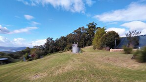Une zone herbeuse avec une petite construction, des arbres et une vue lointaine sur des collines.