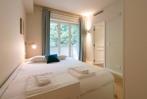 A neatly made bed with white linens and two folded towels, a window with curtains, and a door leading to another room.