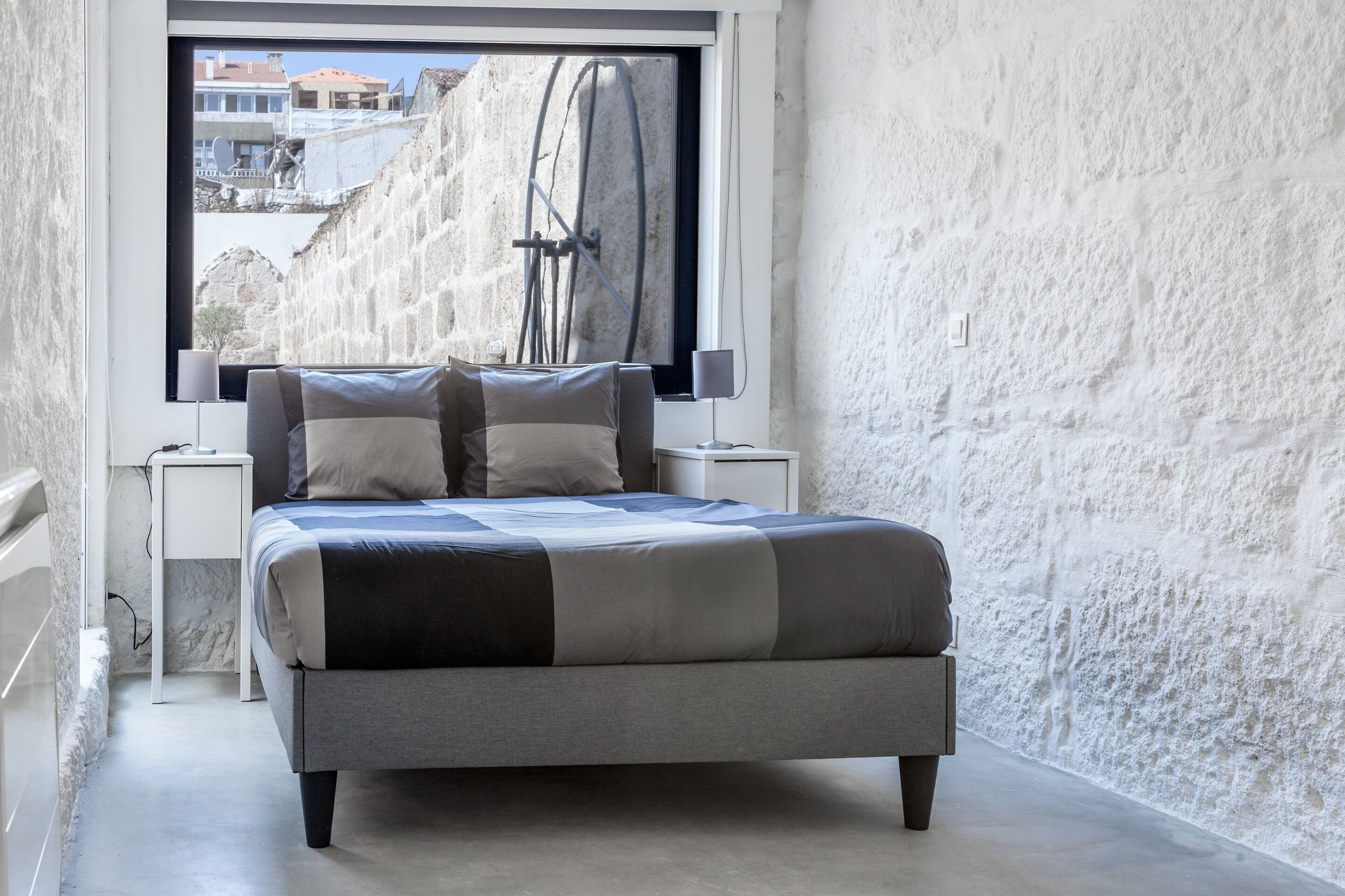 Ein Schlafzimmer mit Bett, Nachttischen, einem Fenster und Blick auf eine Steinmauer und Gebäude.