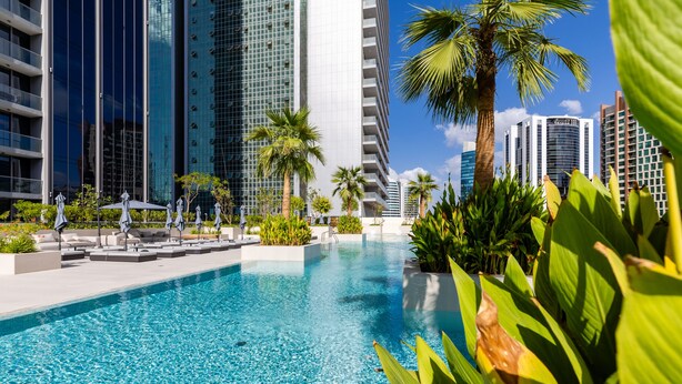 A swimming pool with lounge chairs and palm trees, surrounded by tall buildings. -  Maison Privee - Exquisite Duplex with Private Patio & Loft Living (Dubai)