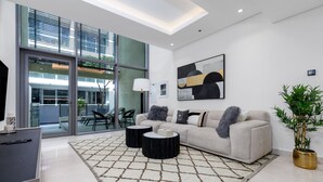 A modern living room with a sofa, two side tables, a large window, and a painting on the wall. -  Maison Privee - Exquisite Duplex with Private Patio & Loft Living (Dubai)