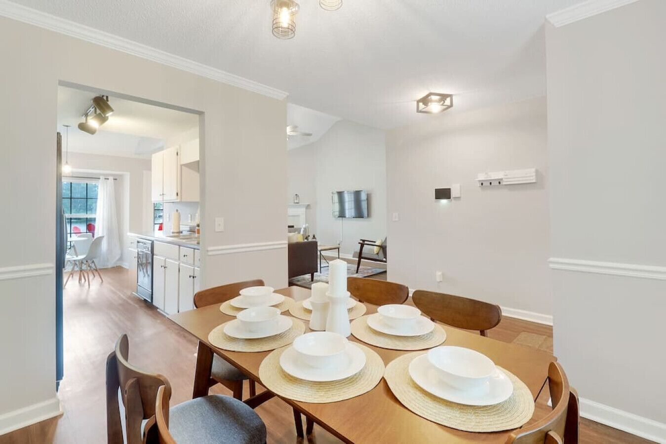 A dining area with a long wooden table set for four, a kitchen in the background, and a TV mounted on the wall.
