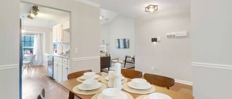 A dining area with a long wooden table set for four, a kitchen in the background, and a TV mounted on the wall.