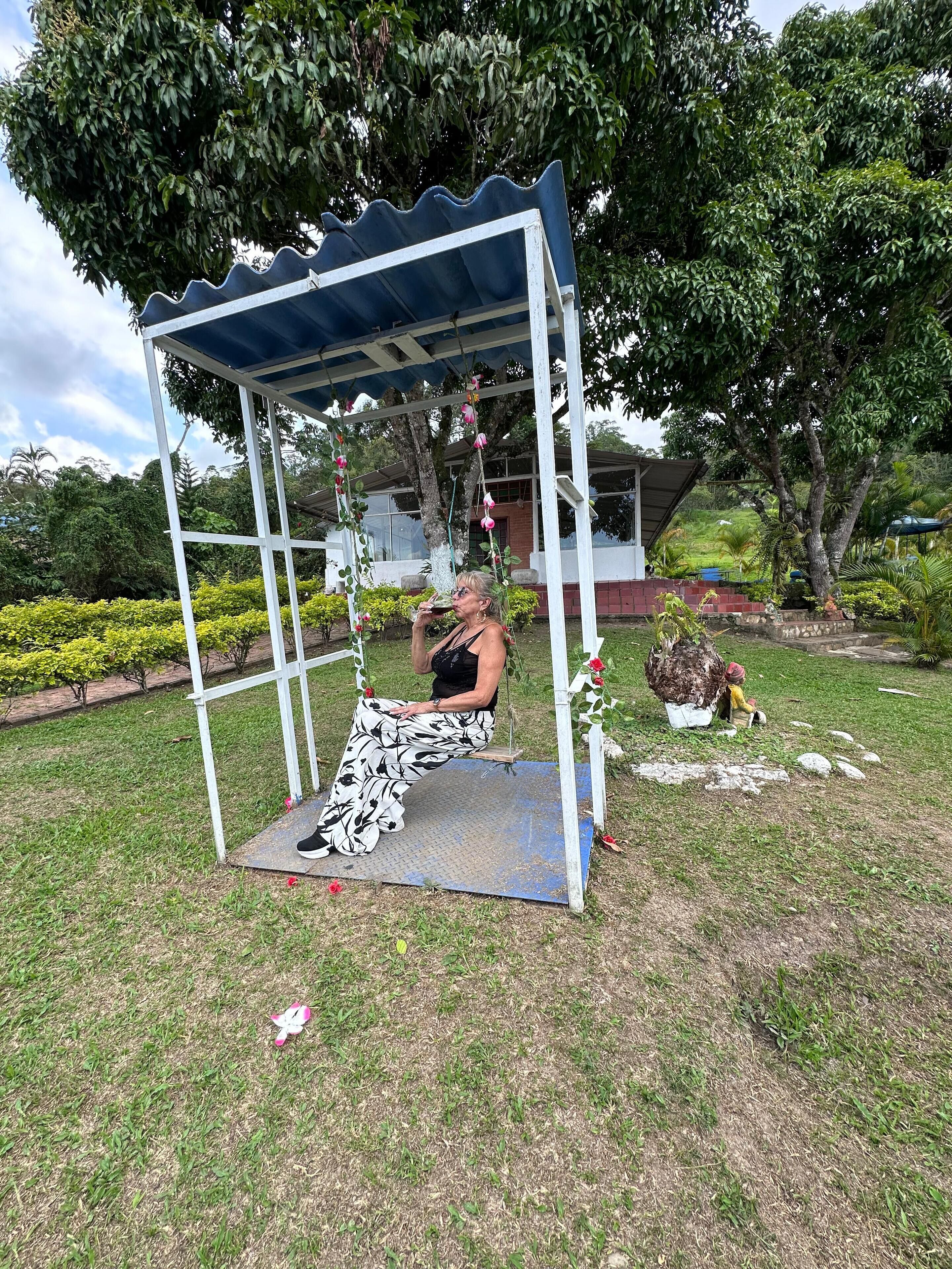 A covered outdoor seating area with a swing, surrounded by greenery and a building in the background.