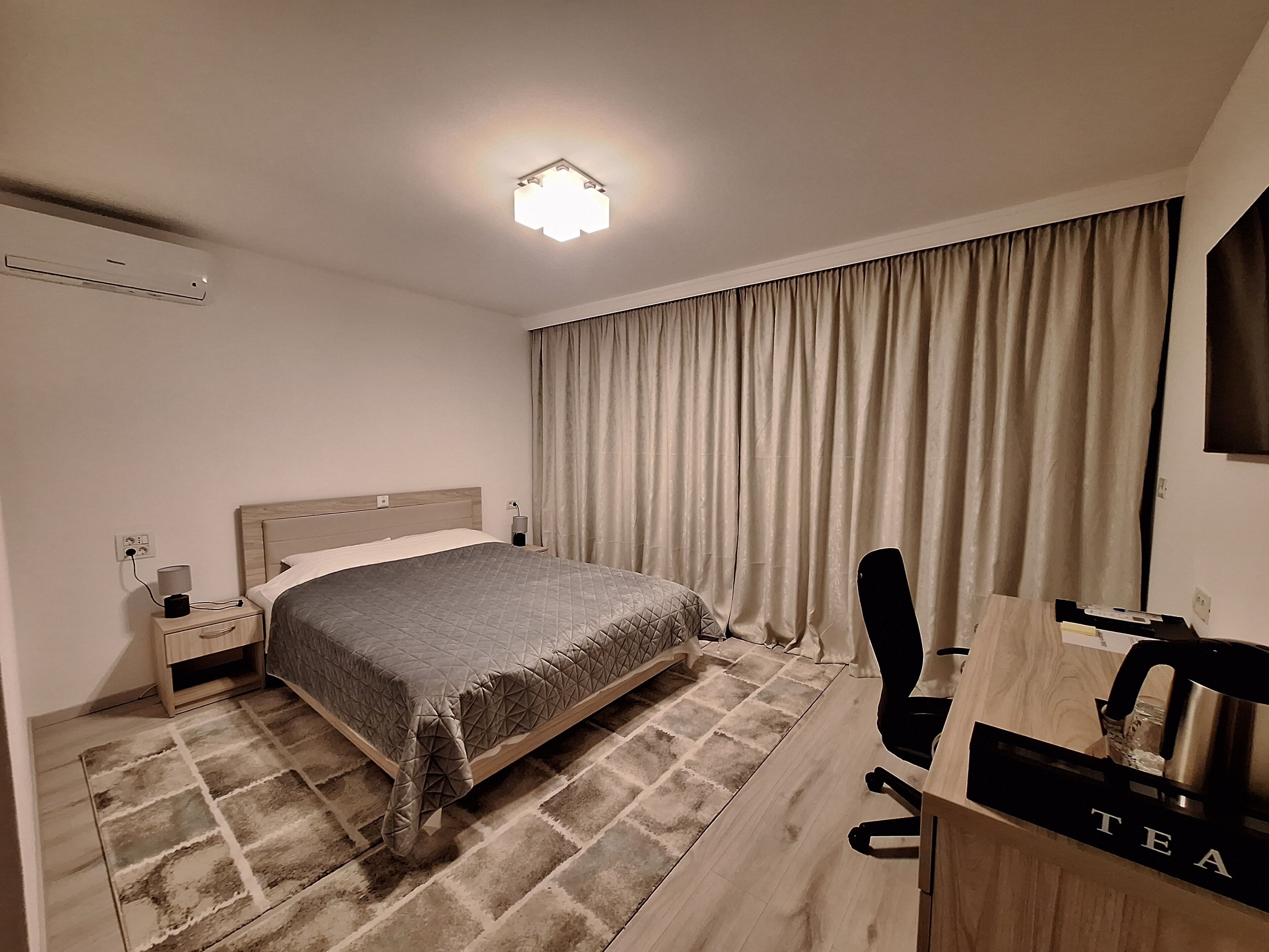 A hotel room with a bed, a desk with a tea station, a chair, a nightstand with a lamp, and a large window with curtains.