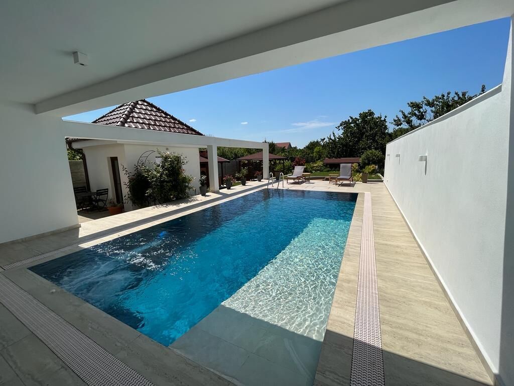 A rectangular swimming pool with clear blue water, surrounded by a white wall and a covered patio area with lounge chairs.