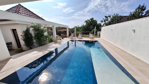 A swimming pool with clear blue water, surrounded by a white wall and a covered area with chairs.