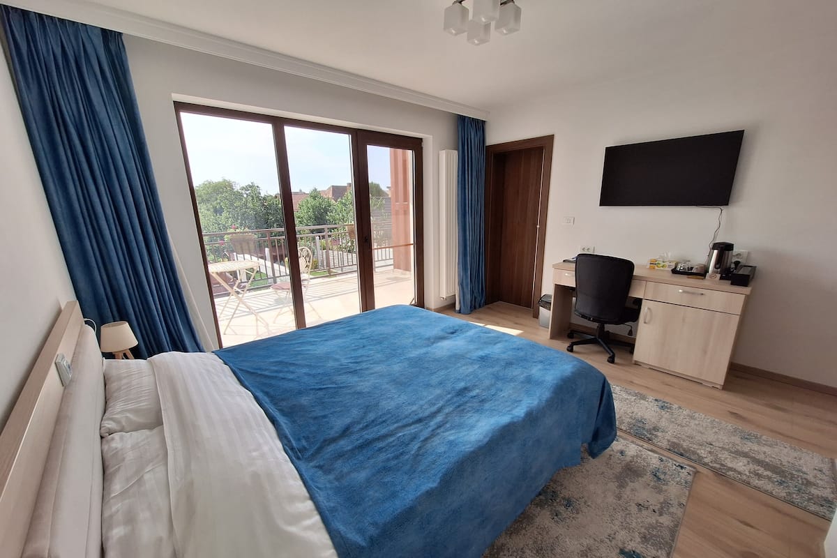 A bedroom with a bed, a desk, a chair, a TV, and a balcony with curtains.