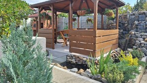 A wooden gazebo with a bench, surrounded by a garden with various plants and a stone wall.