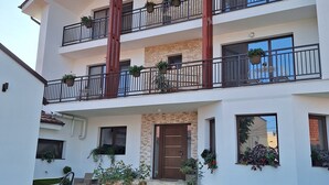 A multi-story building with balconies, a front door, and a garden area with flowers and plants.