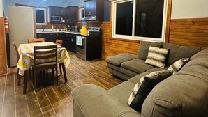 En hyggelig stue med sofa og spisebord, et køkken i baggrunden, træbjælker i loftet og en aircondition.