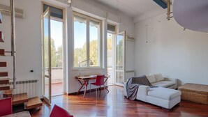 Un salón amplio con suelo de madera, un sofá blanco, una mesa de comedor con sillas y ventanas grandes que ofrecen vistas al exterior.