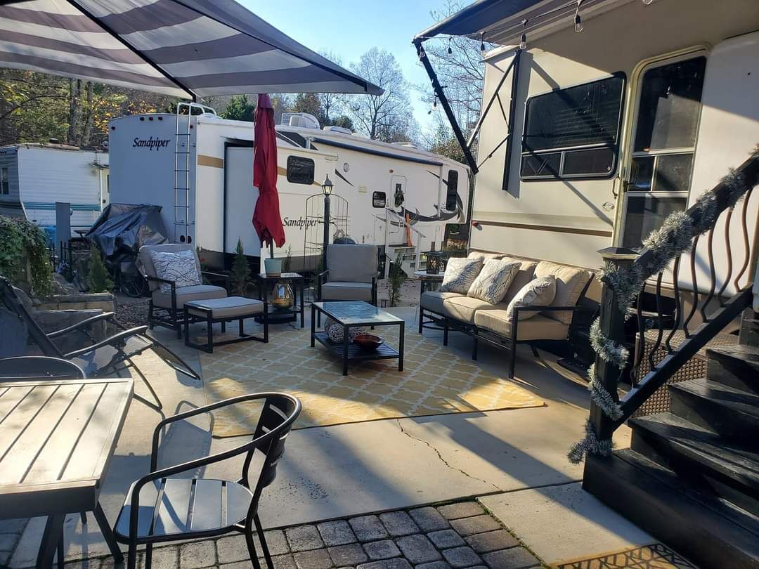 Une terrasse extérieure couverte, aménagée avec un canapé, des fauteuils et une table. Un camping-car est garé à proximité.