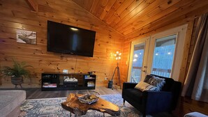 A cozy log cabin interior with a large flat-screen TV, a wooden coffee table, a patterned rug, a sofa, and a potted plant.