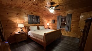 A wooden cabin bedroom with a bed, bedside lamps, a ceiling fan, and a window with curtains.