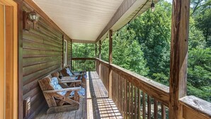 A wooden deck with a railing, a covered porch, and a view of a forested area.