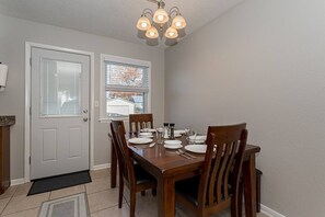 Comedor con una mesa de madera dispuesta para cuatro personas, flanqueada por dos ventanas con persianas, una puerta y una lámpara en el techo.