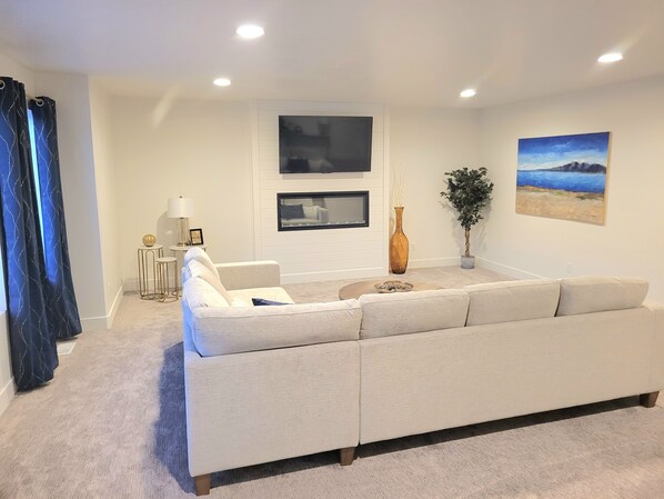 Een moderne woonkamer met een beige bank, een flatscreen televisie aan de muur, een hoge vaas en een ingelijst landschapsbeeld.