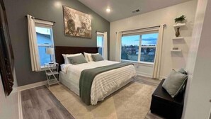 A modern bedroom with a large bed, a nightstand, a desk, and a view of the outdoors through a window.