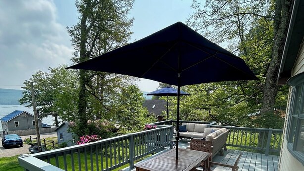 A wooden deck with a table and chairs, a large umbrella, and a view of the lake. - Cozy Hillside Hideaway on Lake Canadarago (Richfield Springs)