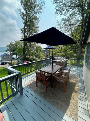A wooden deck with a table and chairs, a large umbrella, and a view of the lake. - Cozy Hillside Hideaway on Lake Canadarago (Richfield Springs)