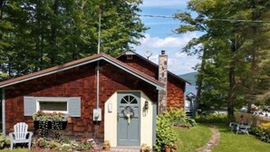 A small, single-story house with a brick exterior, a front door with a window above it, and a small porch with a chair. - Cozy Hillside Hideaway on Lake Canadarago (Richfield Springs)