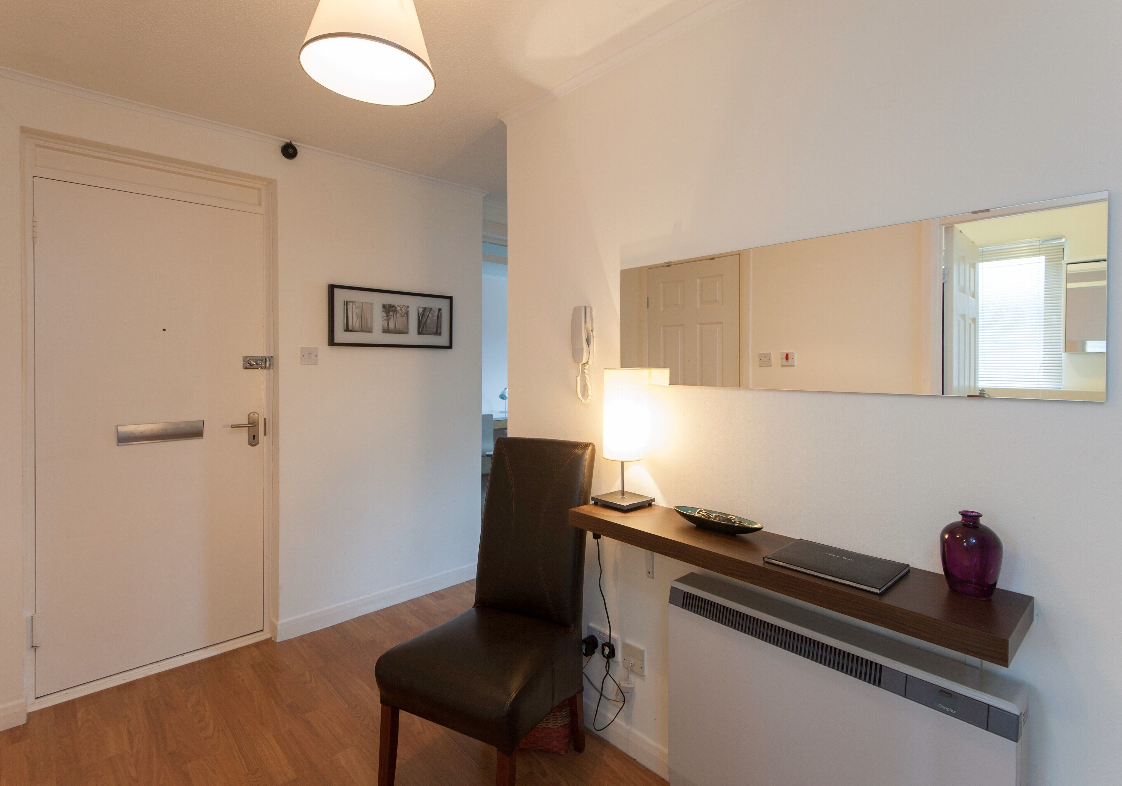 Une pièce avec une porte, une chaise, une table avec une lampe, un miroir et un radiateur.