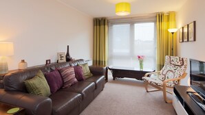 A living room with a brown leather sofa, a wooden chair, a table, a lamp, and a window with curtains. - A modern 2-bedroom apartment in a quiet neighbourhood . Sleeps 4 (Edinburgh)