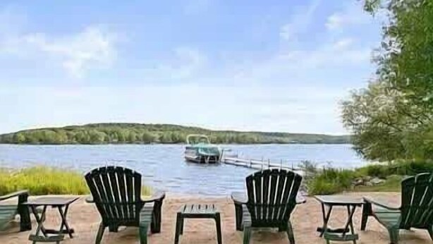 A lakeside seating area with chairs and tables, a boat on the water, and a forested hill in the background. - Lakefront Retreat on Intermediate Lake | Sleeps 14 • Firepit Dogs (Central Lake)