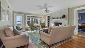 A living room with a beige sofa set, a coffee table, and a fireplace. There are two armchairs and a small table. The room has wooden flooring and a ceiling fan. - Lakefront Retreat on Intermediate Lake | Sleeps 14 • Firepit Dogs (Central Lake)