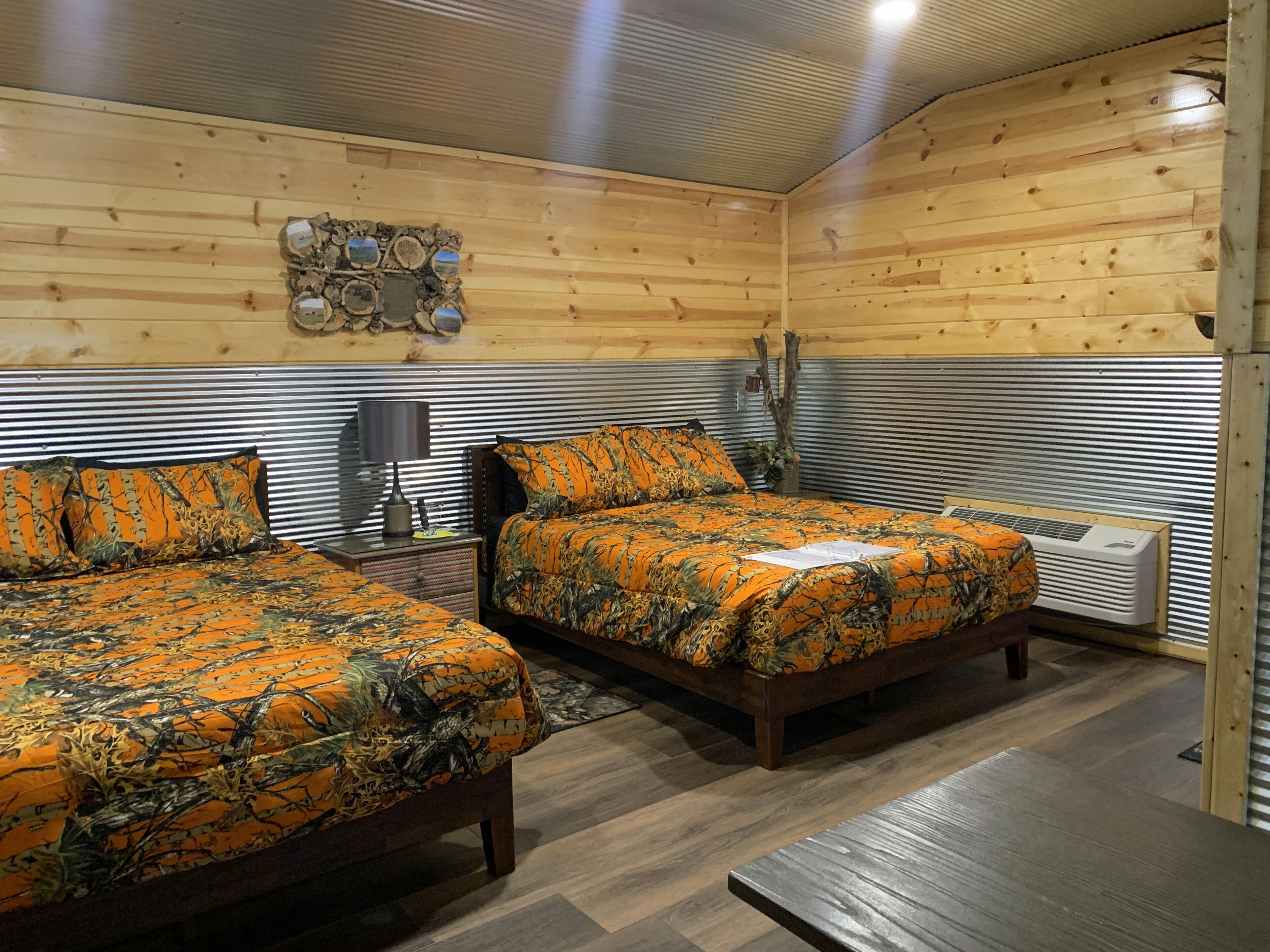 Een kamer met twee houten bedden, een hoofdeinde van metaal, een wandheater en een klein tafeltje met een lamp.