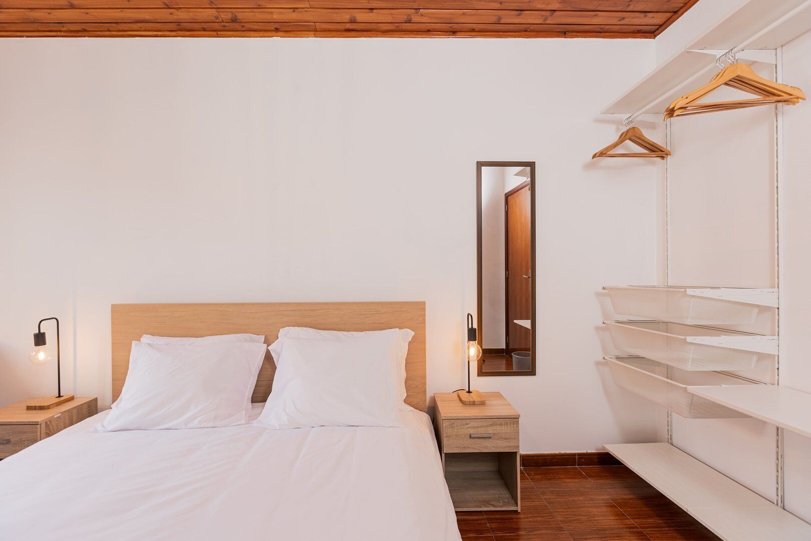 Een slaapkamer met een houten hoofdbord, een nachtkastje met een lamp, een spiegel en een inbouwkast met houten kledinghangers.