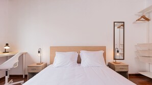 Una camera da letto con una testiera in legno, due cuscini bianchi, una scrivania con una sedia, un comodino con una lampada e uno specchio appeso al muro.
