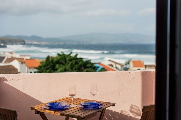 Une table avec deux verres à vin et des assiettes bleues, placée sur un balcon donnant sur l’océan.