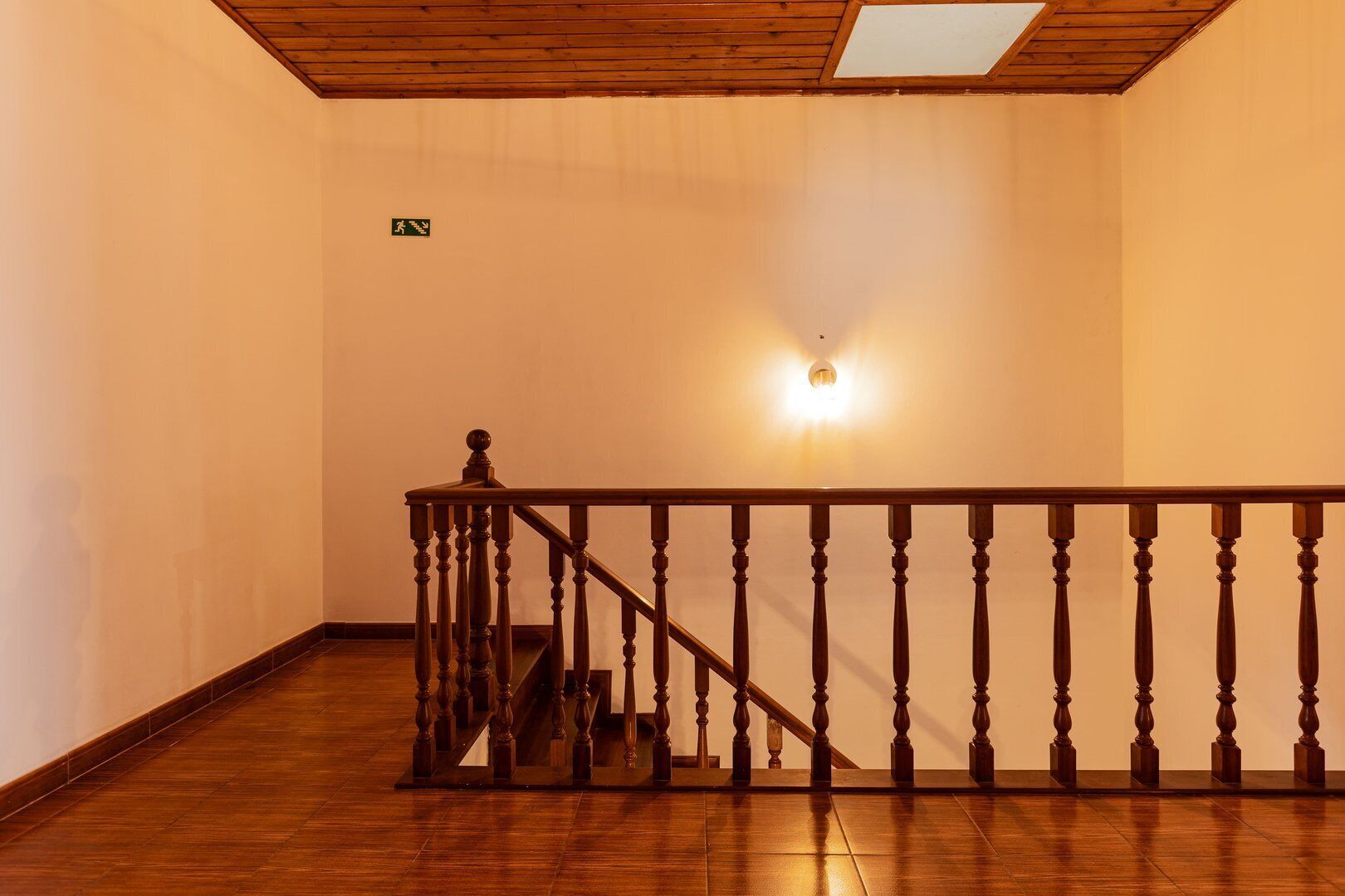 A wooden staircase with a handrail, a light fixture on the wall, and a green exit sign.