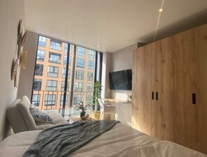 A modern bedroom with a large bed, a wooden wardrobe, a TV mounted on the wall, and a large window offering a view of a building. - Chic Zona Rosa Loft · Rooftop & Coworking (Bogotá)
