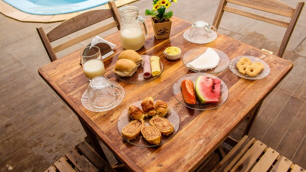 A wooden table with breakfast items including bread, pastries, and a watermelon slice, set outdoors near a pool. - Pousada Barra Grande (Maragogi)