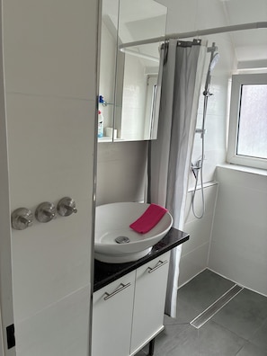 Une salle de bain avec un lavabo blanc, une douche, une fenêtre et un miroir.
