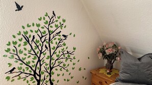 Un dormitorio con techo inclinado, una cama con una manta gris, una mesita de noche de madera con un jarrón de flores y un adhesivo de pared con un árbol y pájaros.