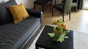 Una sala moderna con un sofá negro, un cojín amarillo, una mesita con un florero, y un comedor con una mesa y sillas.