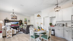 Una cocina moderna con armarios blancos, un comedor con una mesa verde y sillas blancas, y una sala de estar con un sofá y televisor.