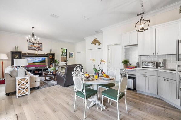Une cuisine moderne avec des meubles blancs, un coin repas avec une table verte et des chaises blanches, et un salon avec un canapé et un téléviseur.
