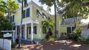 Uma casa amarela de dois andares com varanda, cercada por plantas tropicais.