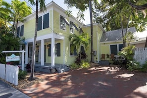 A two-story yellow house with a porch, surrounded by palm trees and a brick pathway.