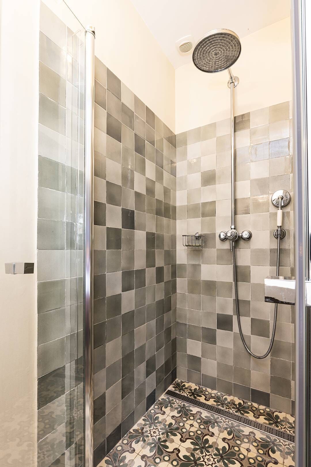 Een douche met glazen deur, betegelde wanden en een patroon vloer.