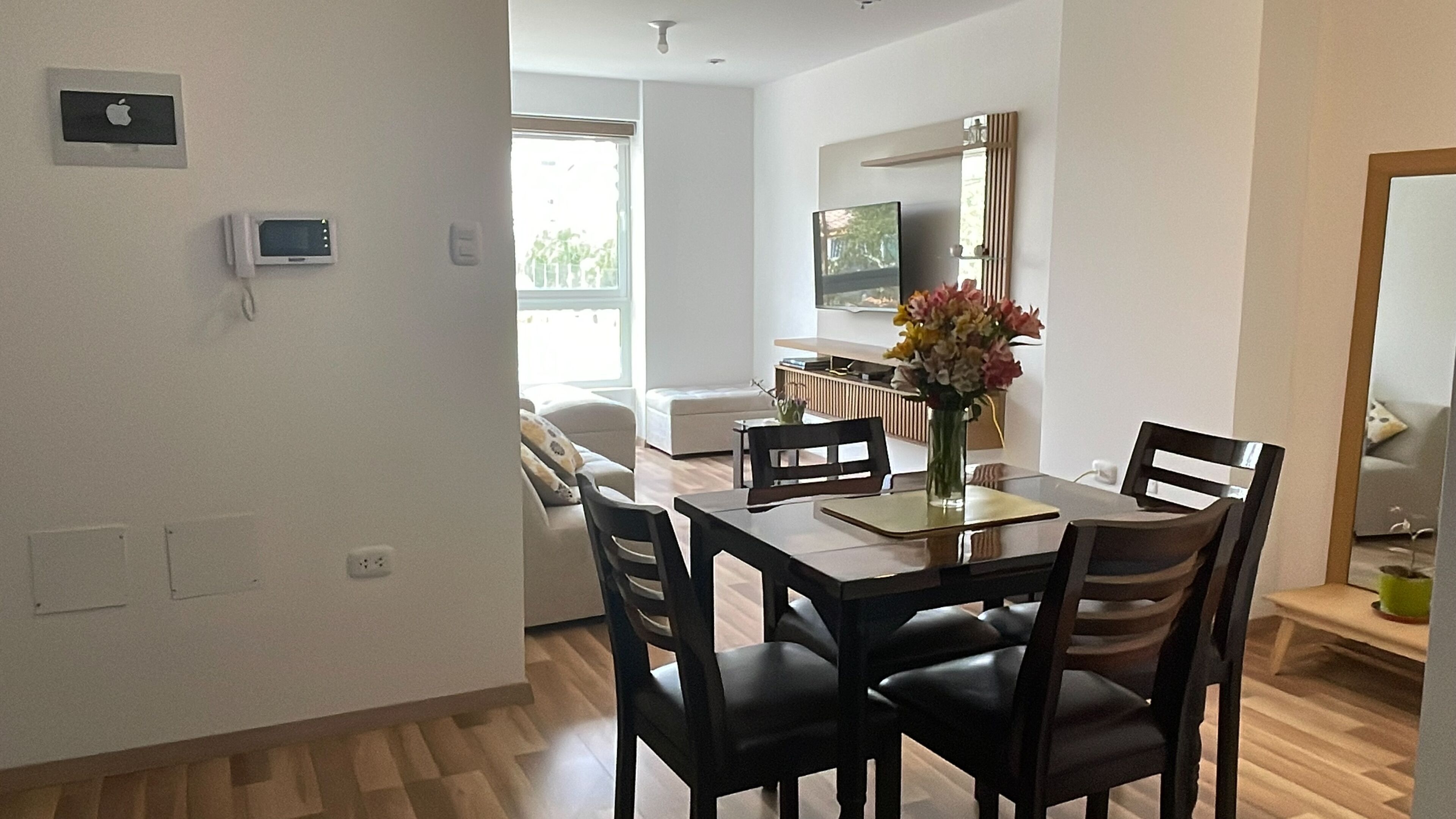 A modern dining area with a wooden table, black chairs, a vase of flowers, and a television in the background.
